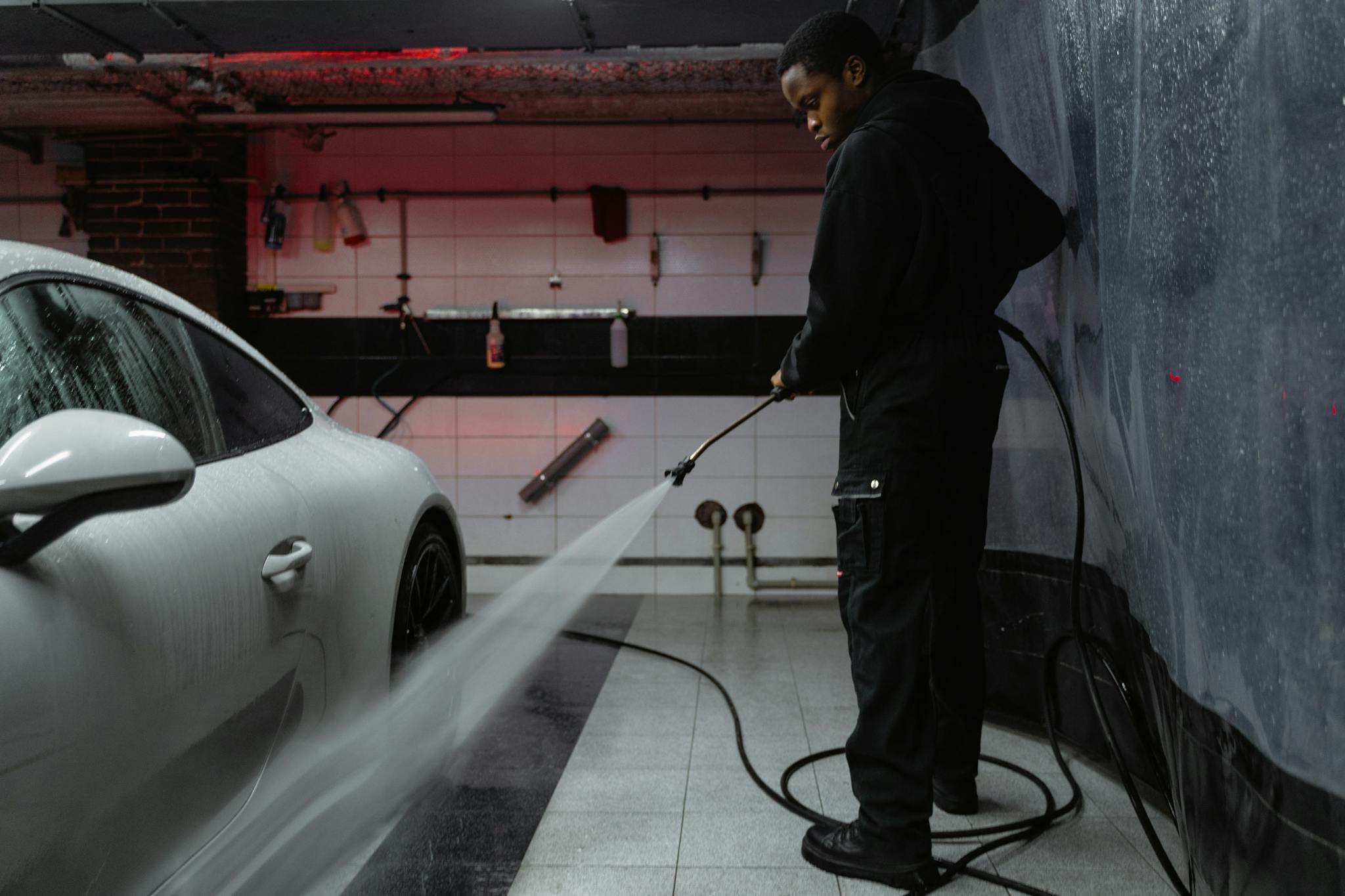African American man cleaning a white car with a pressure hose in an indoor garage setting.